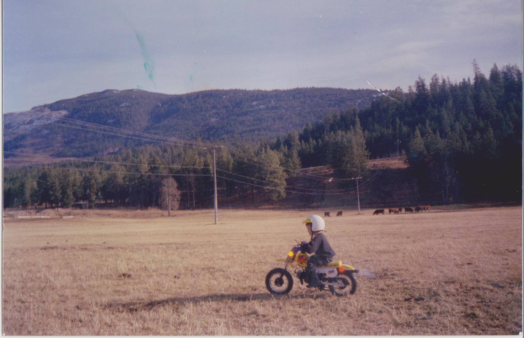 Billy as a young boy in a white helmet, riding his first dirt bike across an open field with mountains in the background.